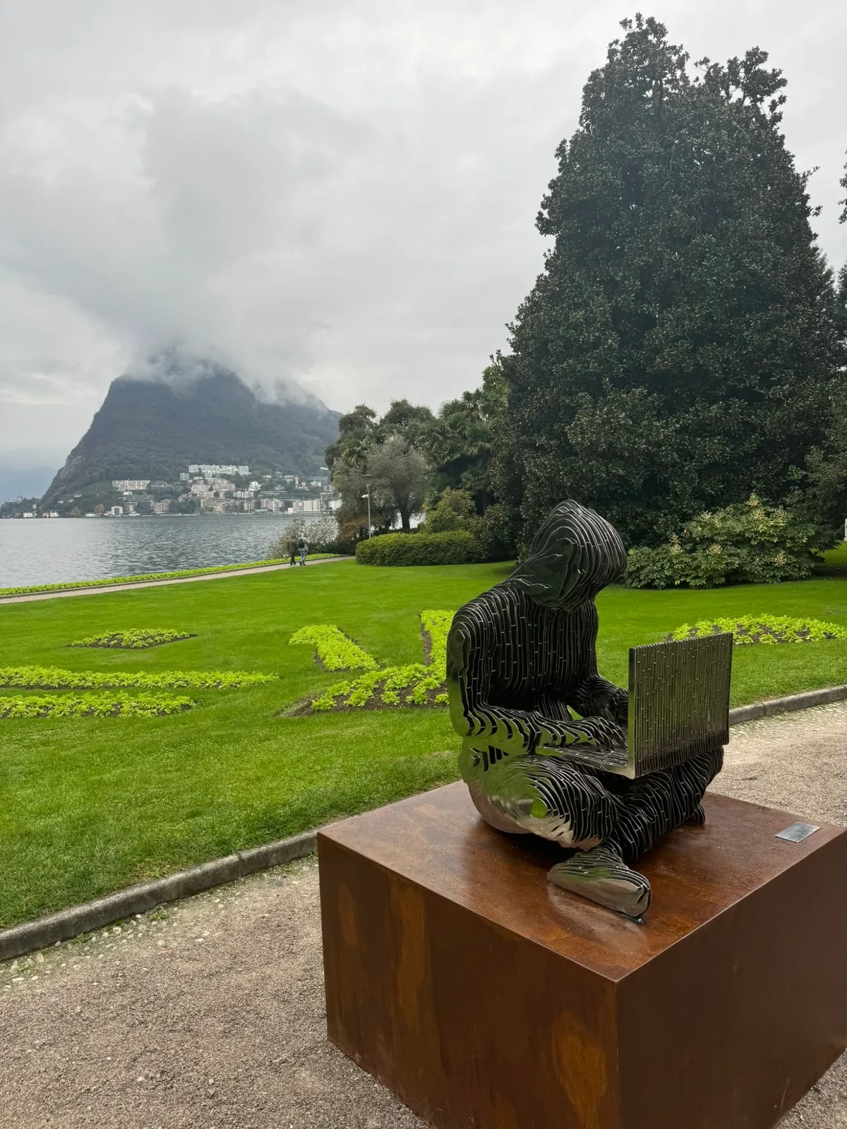 A weathered steel statue of a hooded, faceless figure representing Satoshi Nakamoto, standing among trees in Parco Ciani beside Lake Lugano.
